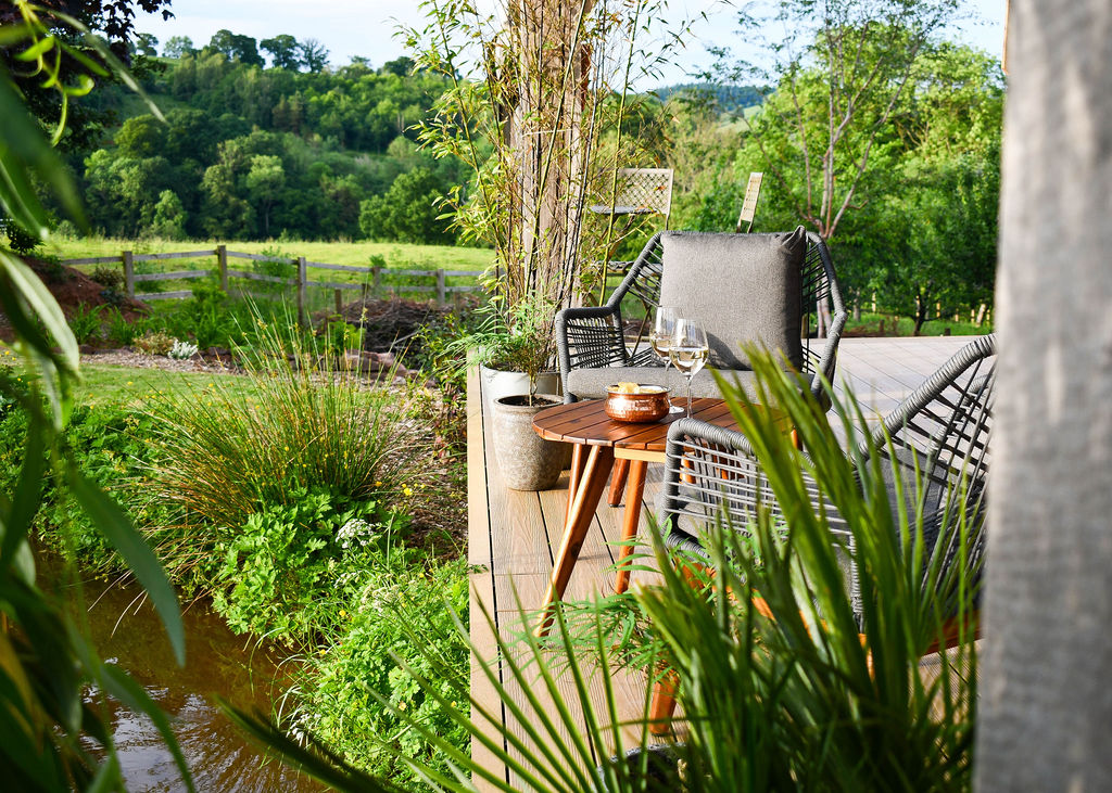 A tranquil outdoor deck at Perry Pond Cabin overlooks lush greenery and a small stream. The area features a comfortable lounge chair with cushions, a side table holding a glass of wine and a small bowl, all shaded by tall plants and surrounded by a serene natural landscape.