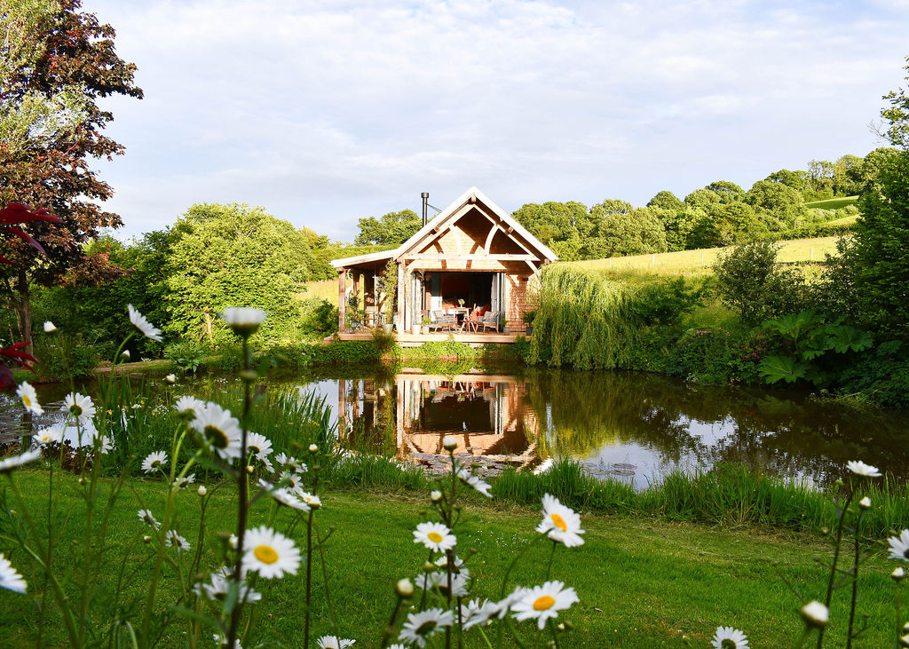 A quaint Pond Cabin with a wooden roof sits beside a serene pond surrounded by lush greenery. The cabin's reflection shimmers in Perry Pond. Tall, blooming daisies in the foreground and rolling hills in the background enhance the idyllic scene.