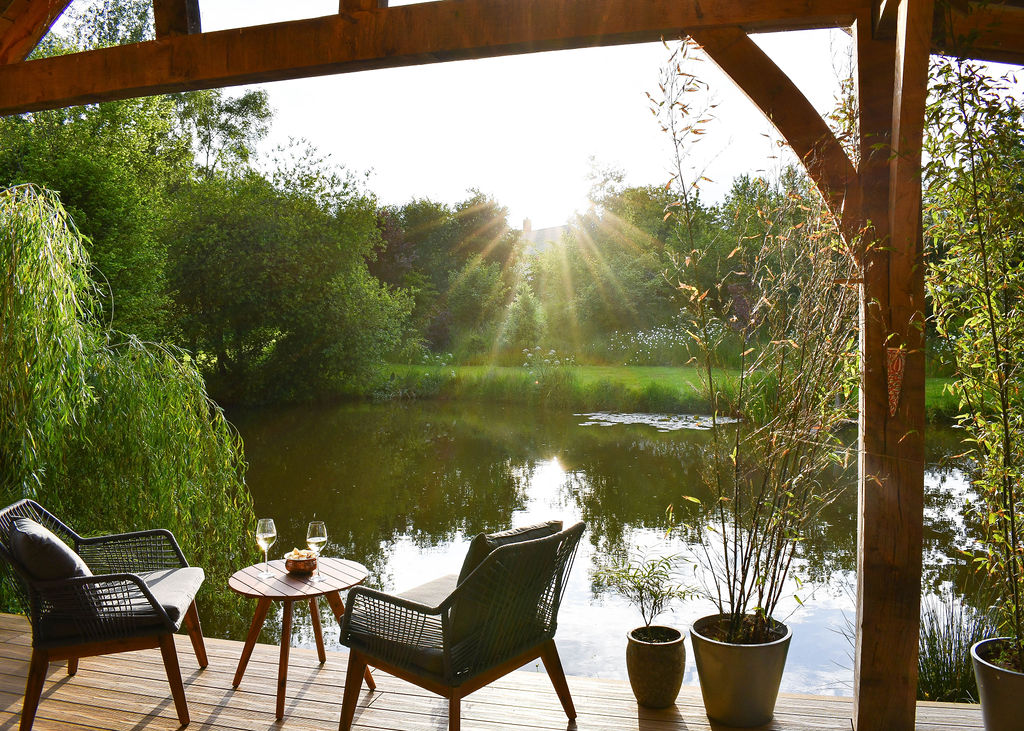 A serene lakeside patio by Perry Pond with two wicker chairs and a small table. On the table are two glasses of wine and a small bowl. The sun is setting over a lush green landscape, creating a warm, golden glow. Nearby, potted plants and trees add to the tranquil atmosphere of this cozy cabin retreat.