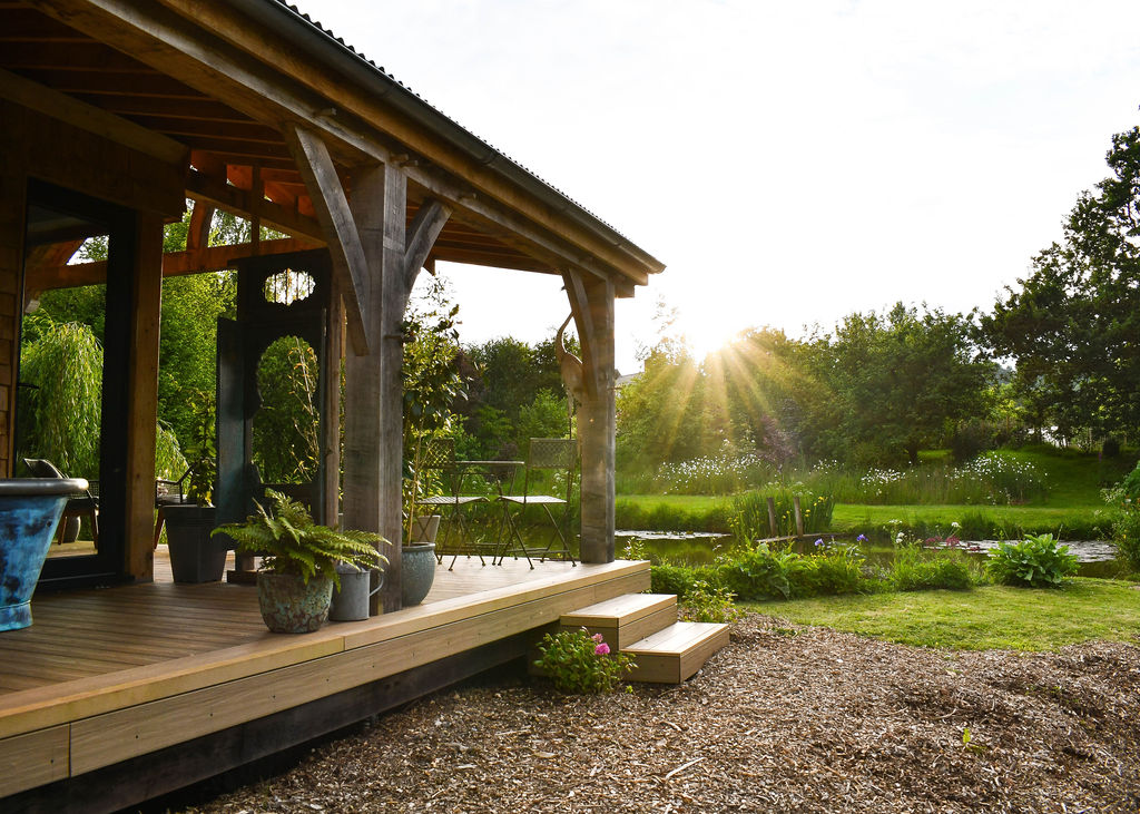 A wooden porch with potted plants leads to a garden with lush greenery and a serene pond known as Perry Pond. Sunlight beams through the trees, creating a warm, inviting atmosphere around the cozy cabin. The scene is tranquil, showcasing nature and a charming outdoor space.