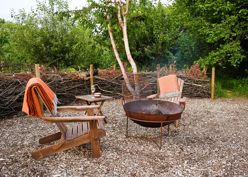 A cozy outdoor seating area at Perry Pond Cabin features two wooden chairs with orange blankets draped over them, facing a large metal fire pit on a gravel ground. A small table between the chairs holds a glass and a candle. The background includes a fence and lush green trees, enhancing the serene ambiance.