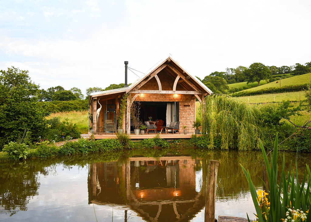 A cozy wooden cabin with a slanted roof, large windows, and a porch sits by the tranquil Perry Pond. Surrounded by lush greenery, rolling hills, and trees in the background, Perry Pond Cabin reflects perfectly in the serene waters of the pond.