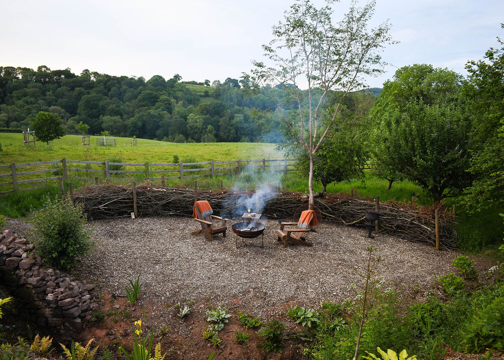 A quaint outdoor seating area with a fire pit surrounded by Adirondack chairs, located in the lush, green garden of Perry Pond Cabin. The area is enclosed by a rustic woven fence, with scenic views of rolling hills and trees in the background. Smoke rises gently from the fire.