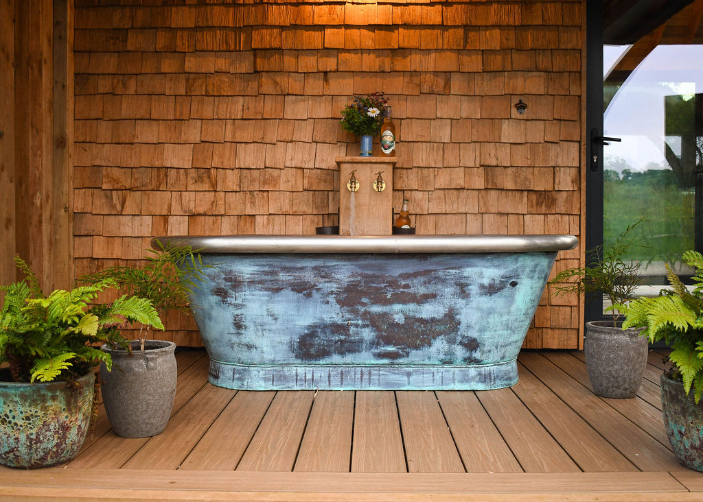 At Perry Pond Cabin, an outdoor wooden deck features a rustic clawfoot bathtub with a weathered blue finish. Behind the tub, a wall of wood shingles is adorned with small potted plants and a decorative faucet. Ferns and other potted plants surround the tub, adding greenery.