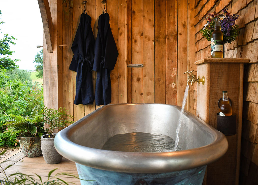 A rustic outdoor bathroom featuring a freestanding metal bathtub with water flowing from a brass faucet. Wooden walls surround the area, with two dark robes hanging on hooks. Plants and a bottle of wine with glasses add to the serene ambiance. A dreamy Devon stay with an outdoor bath. 