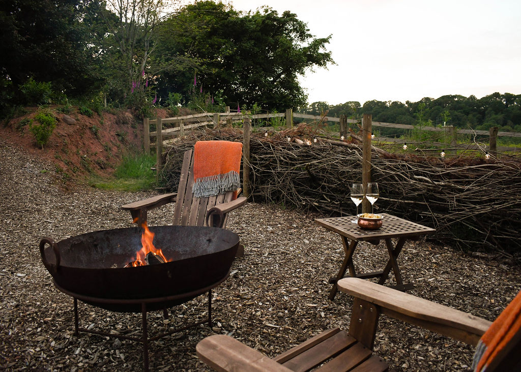 Two wooden chairs with orange blankets are placed around a lit fire pit in a rustic outdoor setting at Perry Pond Cabin. A small table holds two glasses and a bottle, suggesting a cozy, relaxed evening. In the background, a woven fence and greenery provide a serene, natural atmosphere.