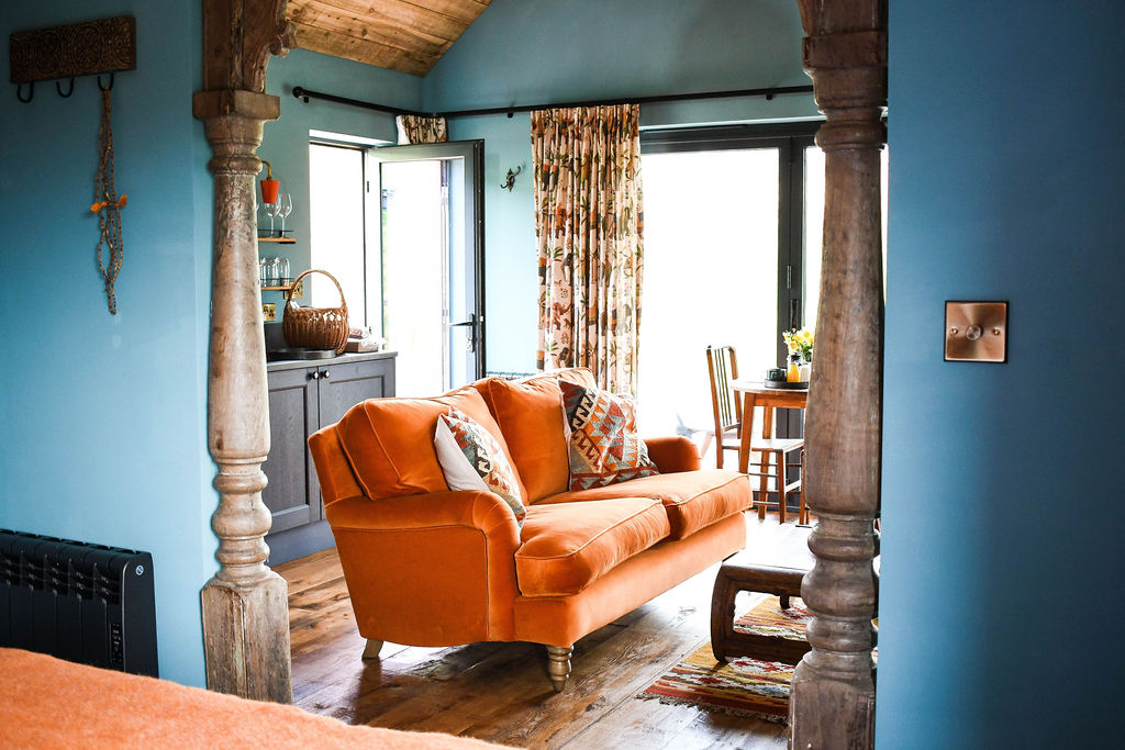 A cozy living area in the Perry Pond Cabin with an orange sofa adorned with patterned cushions, rustic wooden columns framing the view. The room features blue walls, a small dining table with chairs by the window, floral curtains, and a glimpse of a kitchen area in the background.