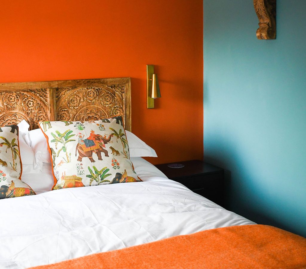 A cozy bedroom at Perry Pond Cabin features a carved wooden headboard, vibrant orange and teal walls, and matching decorative pillows with botanical and animal prints. A wicker lampshade hangs above, while a wall-mounted shelf with books adorns the teal wall. An orange throw decorates the bed.