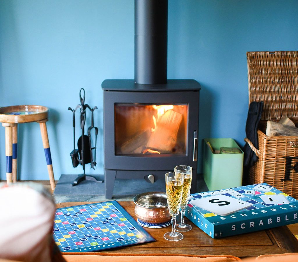 A cozy living room scene in a cabin by Perry Pond features a warm, lit fireplace against a blue wall. A Scrabble game board is set up on a wooden table with the box nearby. Two glasses of bubbly beverages, a small bowl, a stool, and a wicker basket are also present.