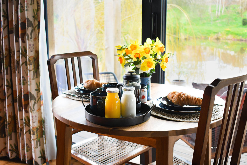 A small round wooden dining table set for two with black plates, croissants, mugs, and jugs of orange juice and milk. A bouquet of yellow flowers is in the center. The table is by a large window with a scenic view of Perry Pond and greenery outside the cabin.