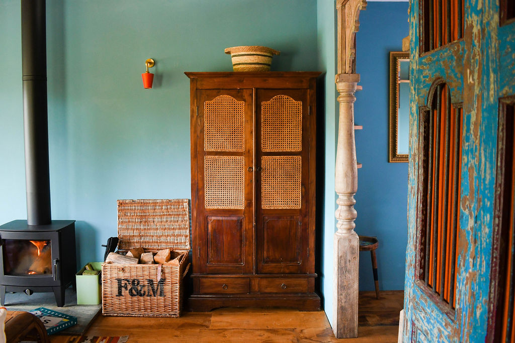 A cozy room in Perry Pond Cabin features a wood-burning stove on the left, a wicker basket filled with firewood, and a vintage wooden wardrobe with cane doors. The walls are painted blue, and a tall pillar stands on the right beside a multicolored distressed door.