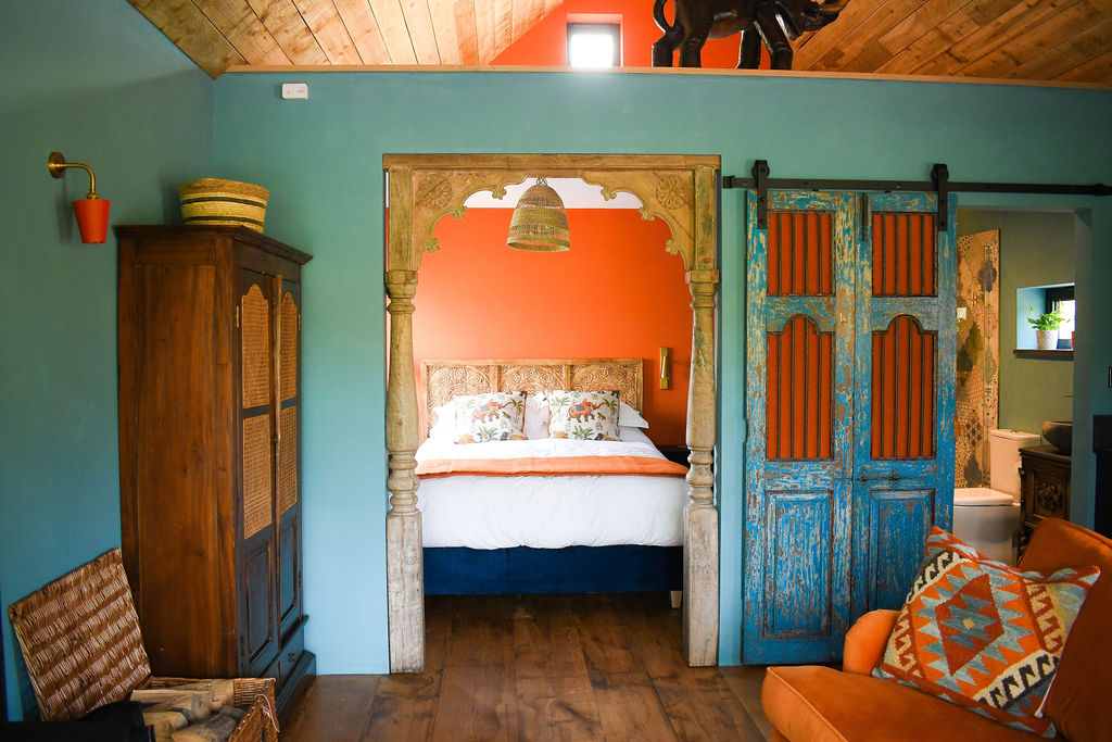 A cozy bedroom at Perry Pond Cabin features vibrant decor: a bed with floral pillows beneath a rustic wooden arch, flanked by teal-blue sliding doors. An orange accent wall complements the interior. A wooden cabinet and orange armchair are placed in the foreground on a wooden floor.