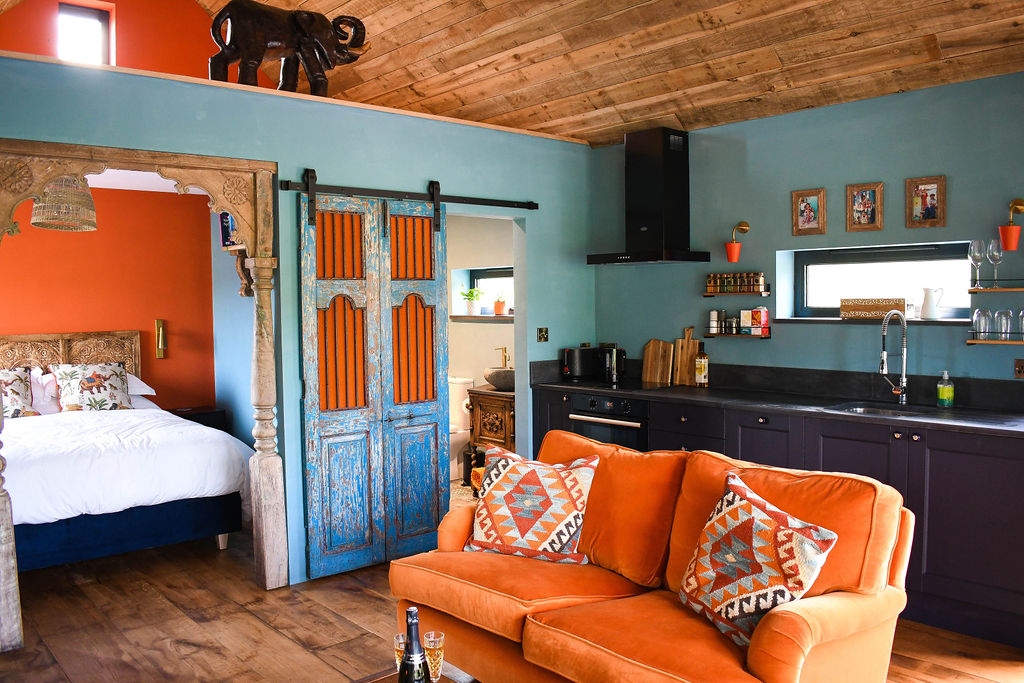 A cozy living space at Perry Pond Cabin features an orange couch with patterned cushions, a kitchen with dark cabinets and blue walls, and a partially hidden bedroom through a rustic sliding door. The wooden ceiling and various decorations, including an elephant figurine, add to the cabin's charm.