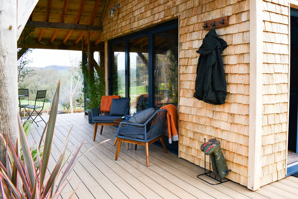 A wooden deck with wicker chairs, orange blankets, and black cushions under a covered patio creates the perfect cabin vibe. A black coat hangs on a hook on the wooden shingle wall, with a small green pouf nearby. In the background, trees frame a distant view of Perry Pond's serene landscape.