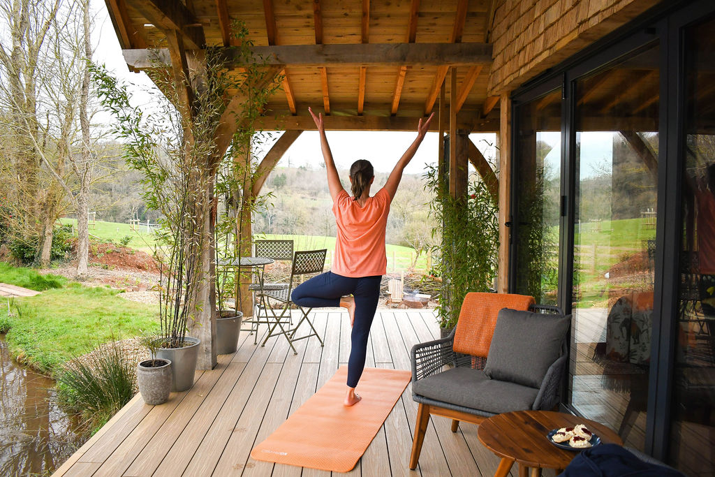 A person stands on an orange yoga mat in a tree pose with arms extended upwards. They are on a wooden deck at Perry Pond Cabin, with outdoor furniture, surrounded by plants and overlooking a scenic landscape of trees and hills. The space is covered by a wooden roof.