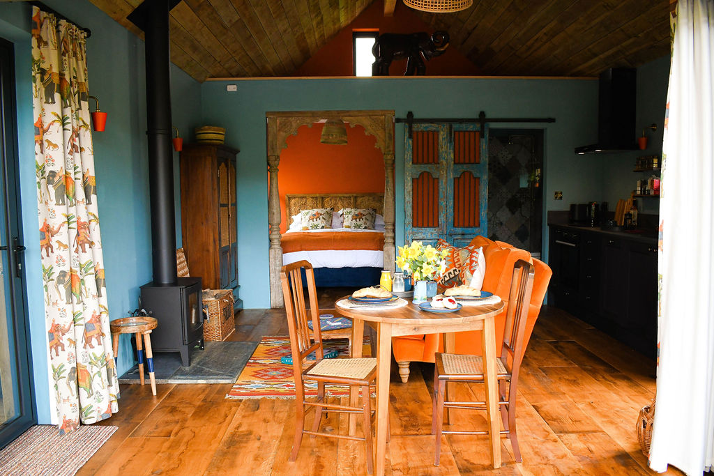 A cozy, colorful interior picture of Perry Pond Cabin features rustic wooden floors, a small dining table with four chairs, a bed adorned with red and white bedding, an array of eclectic furniture, and vibrant decor including floral curtains and an ornate wooden arch.