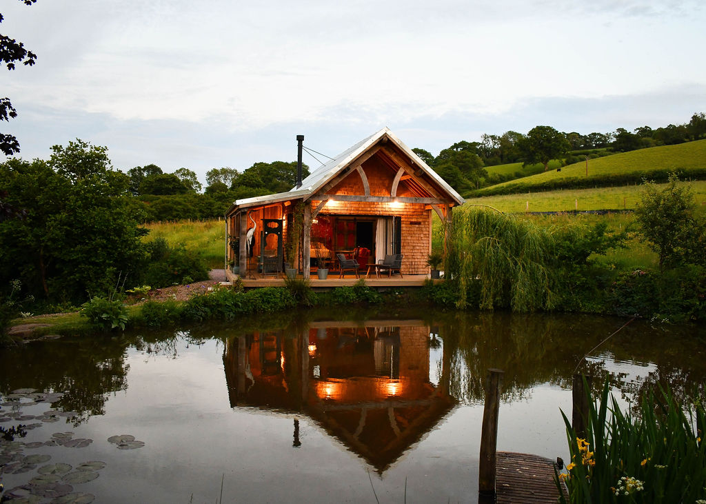 Perry Pond Cabin is a cozy wooden retreat with bright interior lights, sitting by a tranquil pond that reflects its clear water. The lush green hills, trees, and grassy area nearby create a serene landscape. A small dock can be seen in the foreground.