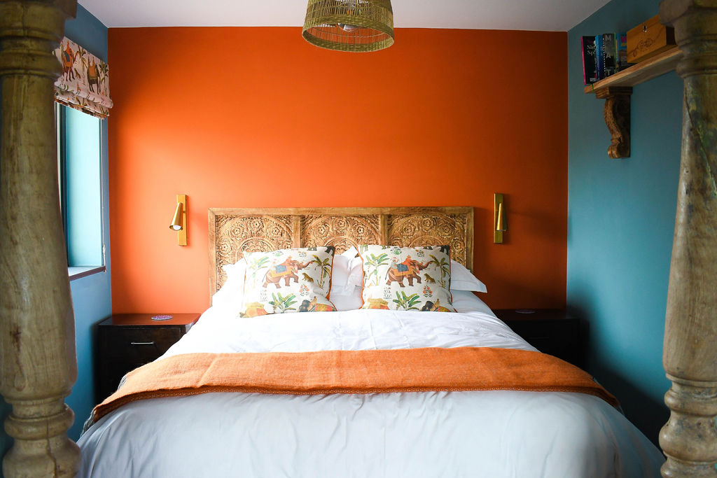 A cozy bedroom in Perry Pond Cabin boasts a vibrant orange accent wall behind a wooden headboard with intricate carvings. The bed has white linens, an orange throw blanket, and decorative pillows featuring an elephant pattern. The room features teal side walls, a window, and a hanging light fixture.