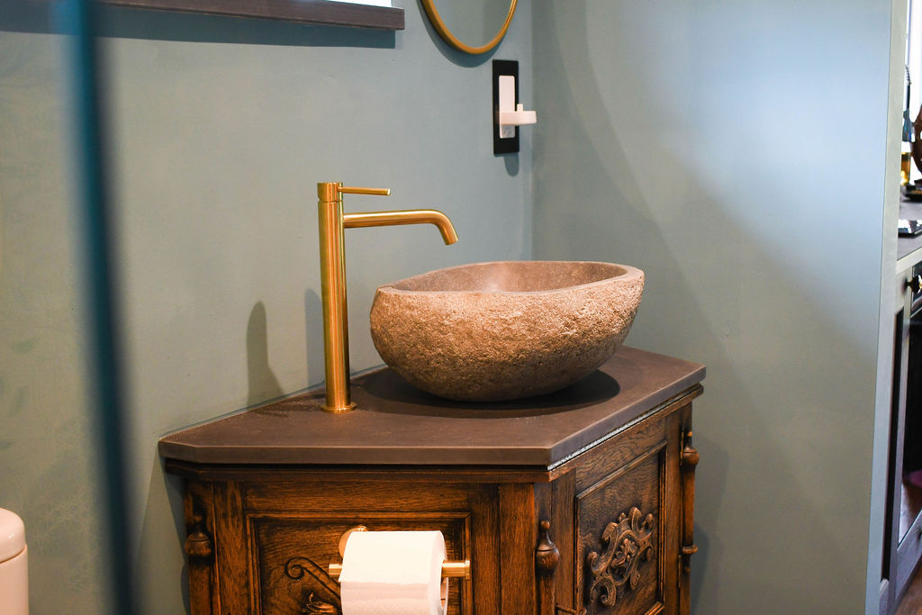 A modern bathroom sink area at Perry Pond Cabin features a stone basin atop a dark wooden vanity with intricate carvings. The faucet is a sleek, straight brass design. A round mirror with a gold frame hangs on the wall, and there's a wall-mounted soap dispenser beside it.