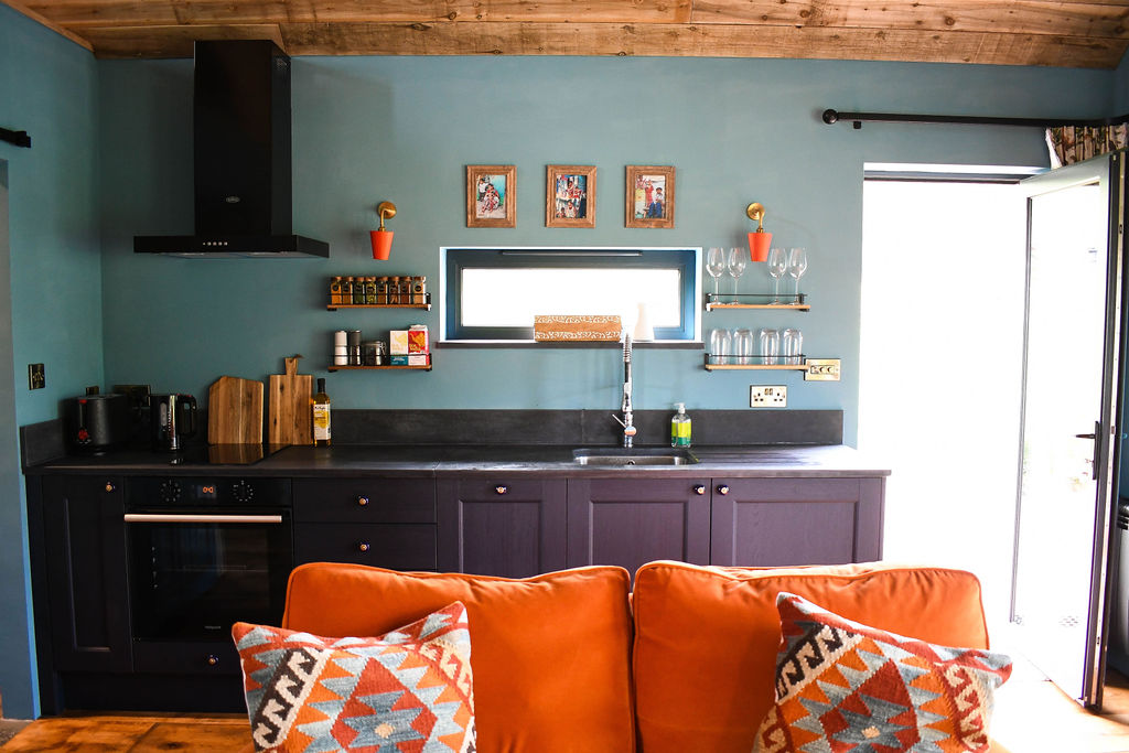 A cozy kitchen with teal walls, dark cabinets, and a wooden counter feels like a charming cabin retreat. Shelves hold spices, glasses, and framed photos. A window above the sink offers natural light and views of Perry Pond. An orange sofa with patterned cushions is in the foreground.