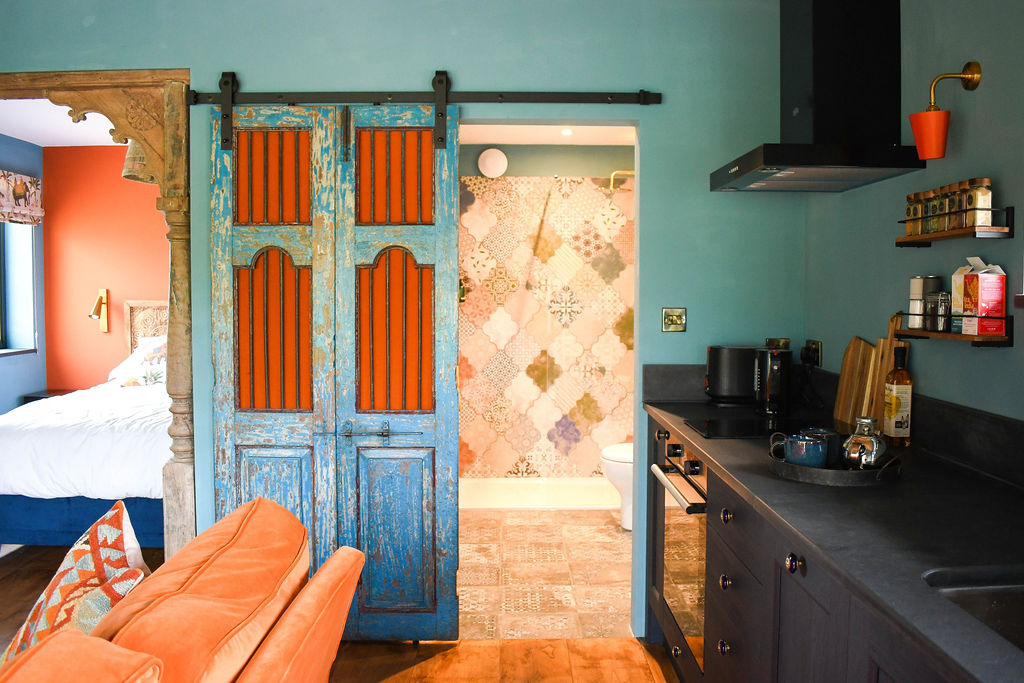 A cozy Perry Pond Cabin-inspired studio apartment with a teal and orange color scheme. The kitchen has dark countertops and modern appliances. Vibrant blue sliding doors lead to a bathroom with patterned tiles. To the left, a bedroom with peach walls and a white bed is partially visible.