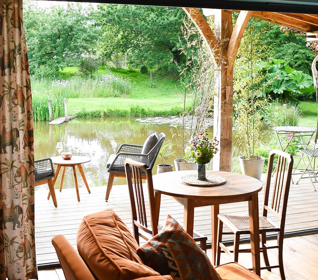 A cozy living area in the Perry Pond Cabin features an orange sofa with colorful cushions, a round wooden dining table, and chairs. Through the large open patio doors, a wooden deck overlooks serene Perry Pond with outdoor seating and lush greenery, evoking a peaceful, nature-filled atmosphere.