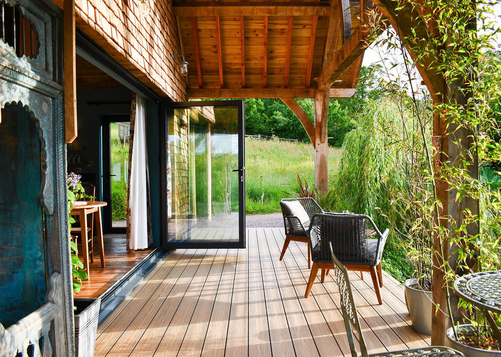 A cozy outdoor deck attached to the Perry Pond Cabin features wicker chairs and a small table. The area is surrounded by lush greenery, including a weeping willow. A sliding glass door opens to the interior, offering a seamless transition between indoors and outdoors.