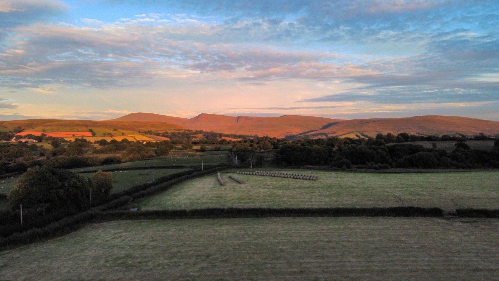 A scenic landscape shows rolling hills bathed in the warm glow of a setting sun. The foreground includes lush green fields with hedgerows and a few scattered trees, reminiscent of Nantseren's tranquil beauty. The sky is filled with soft clouds, creating a serene and peaceful atmosphere.