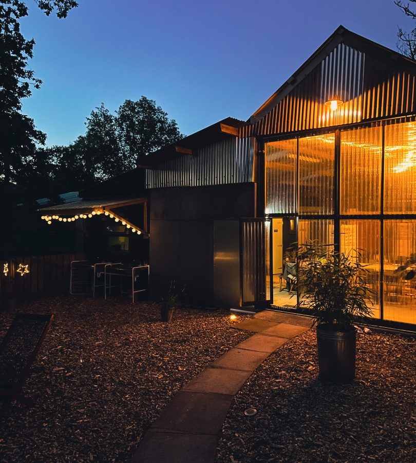 A cozy, illuminated modern cabin with large windows and a metal exterior is seen at dusk. String lights hang outside, casting a warm glow over the Nantseren path lined with rocks and plants leading to the entrance. Trees frame the night sky in the background.