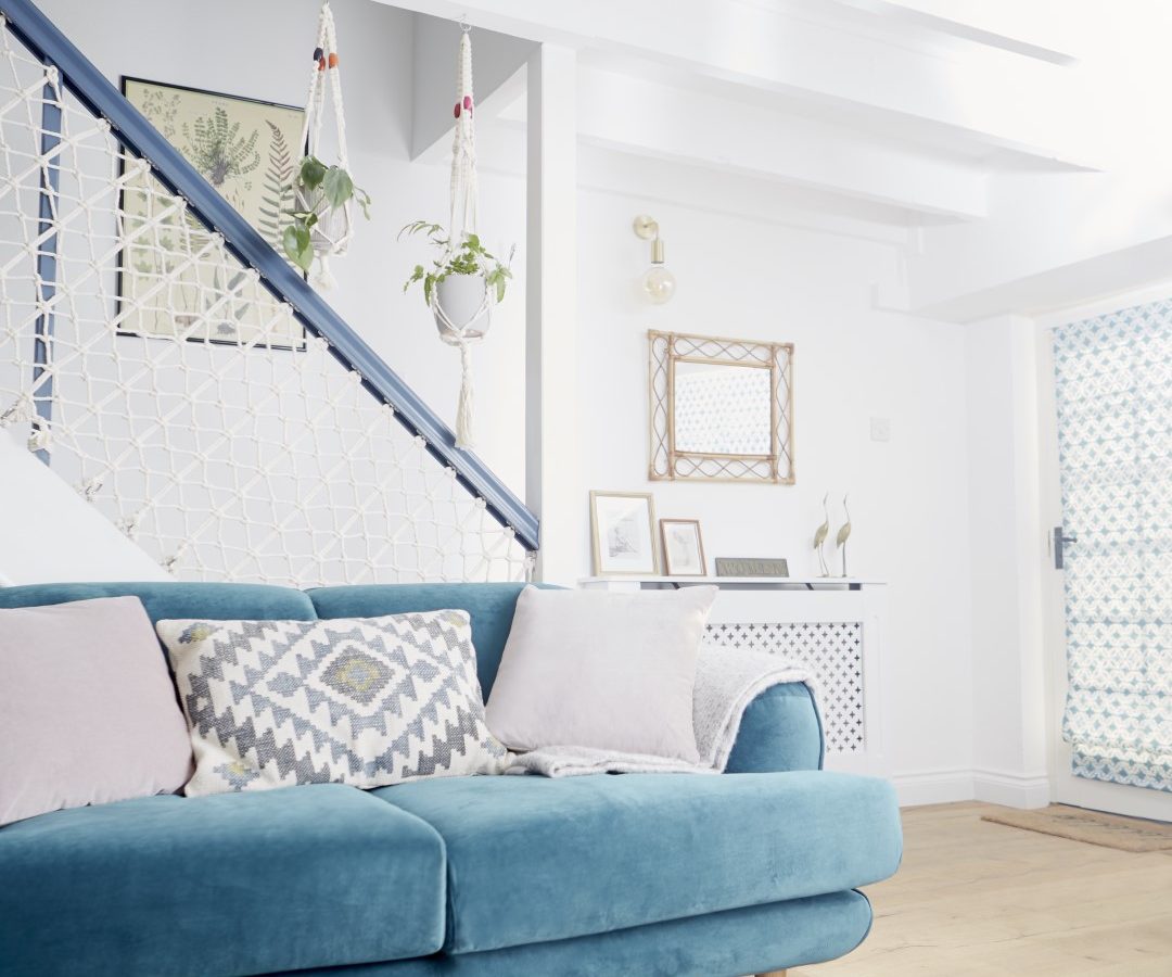 A bright living room in Poachers Pocket Cottage features a light blue sofa adorned with patterned cushions. The white-walled, wooden-floored space boasts a macramé hammock plant holder on the staircase railing. The room includes a blue-and-white geometric door, framed photos, and decor on white furniture.