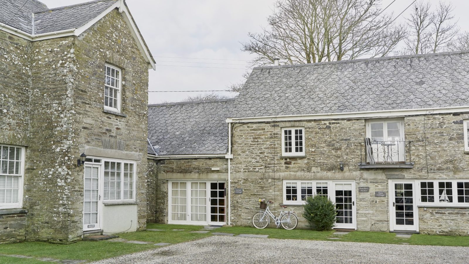 Poachers Pocket Cottage is a charming stone building with multiple sections featuring wooden doors and white-framed windows. A white bicycle leans against the wall near a small bush, while a gravel pathway winds in the foreground, framed by leafless trees in the background.