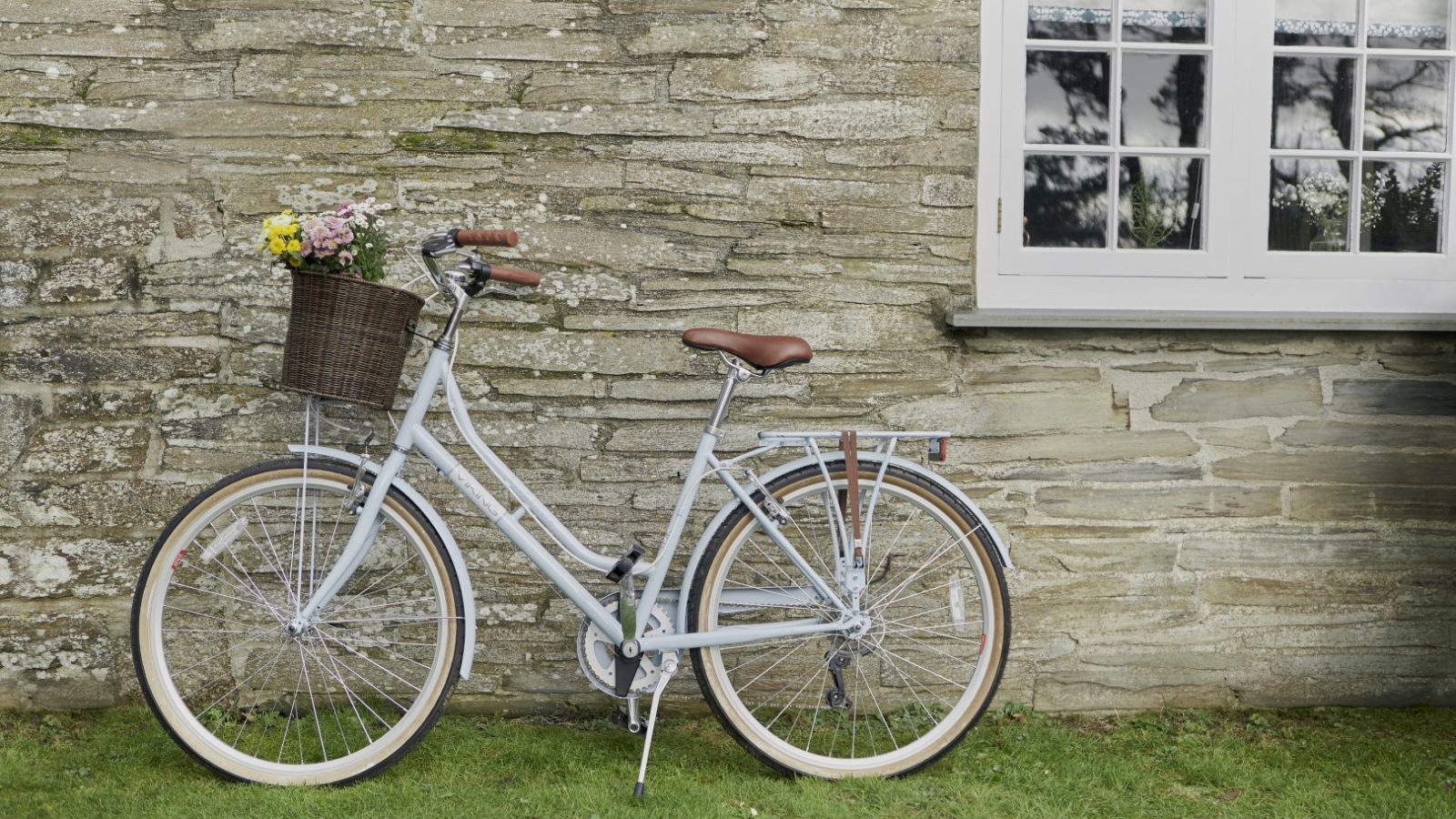 A light gray bicycle with a brown leather seat rests against the weathered stone wall of Poachers Pocket Cottage. A woven basket with a bouquet of colorful flowers is attached to the front, beneath a white-framed window. The bike stands on a patch of green grass.