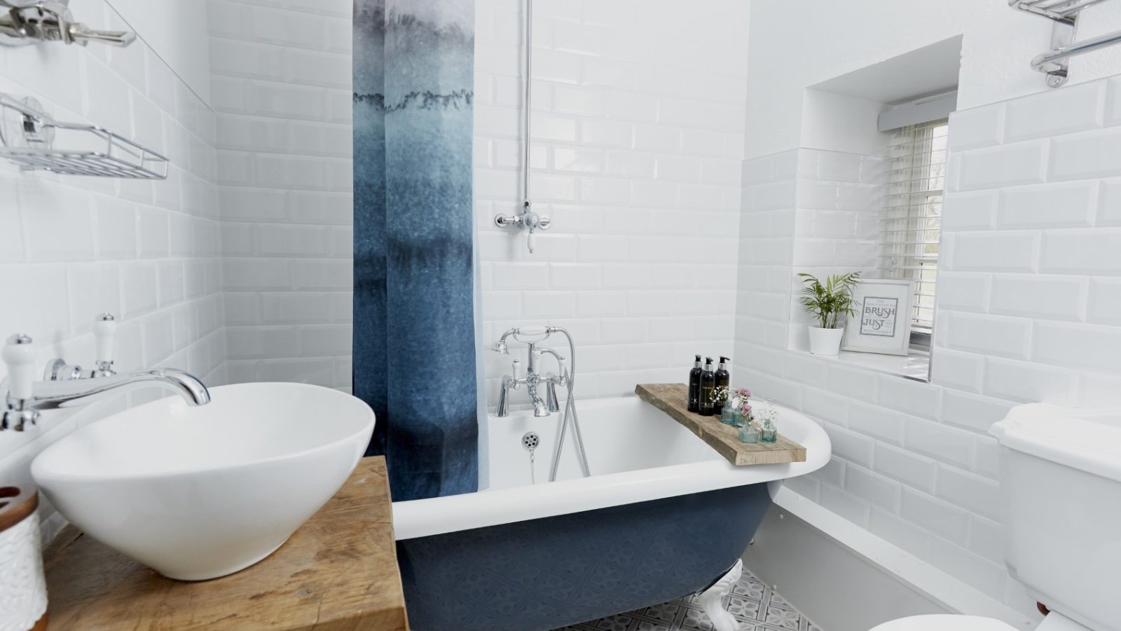 A modern bathroom with white tiled walls and a decorative patterned tile floor. It features a clawfoot bathtub with a wooden tray, reminiscent of cottage charm, a white ceramic sink on a wooden countertop, and a gradient blue shower curtain. A potted plant, framed picture, and window add decor.