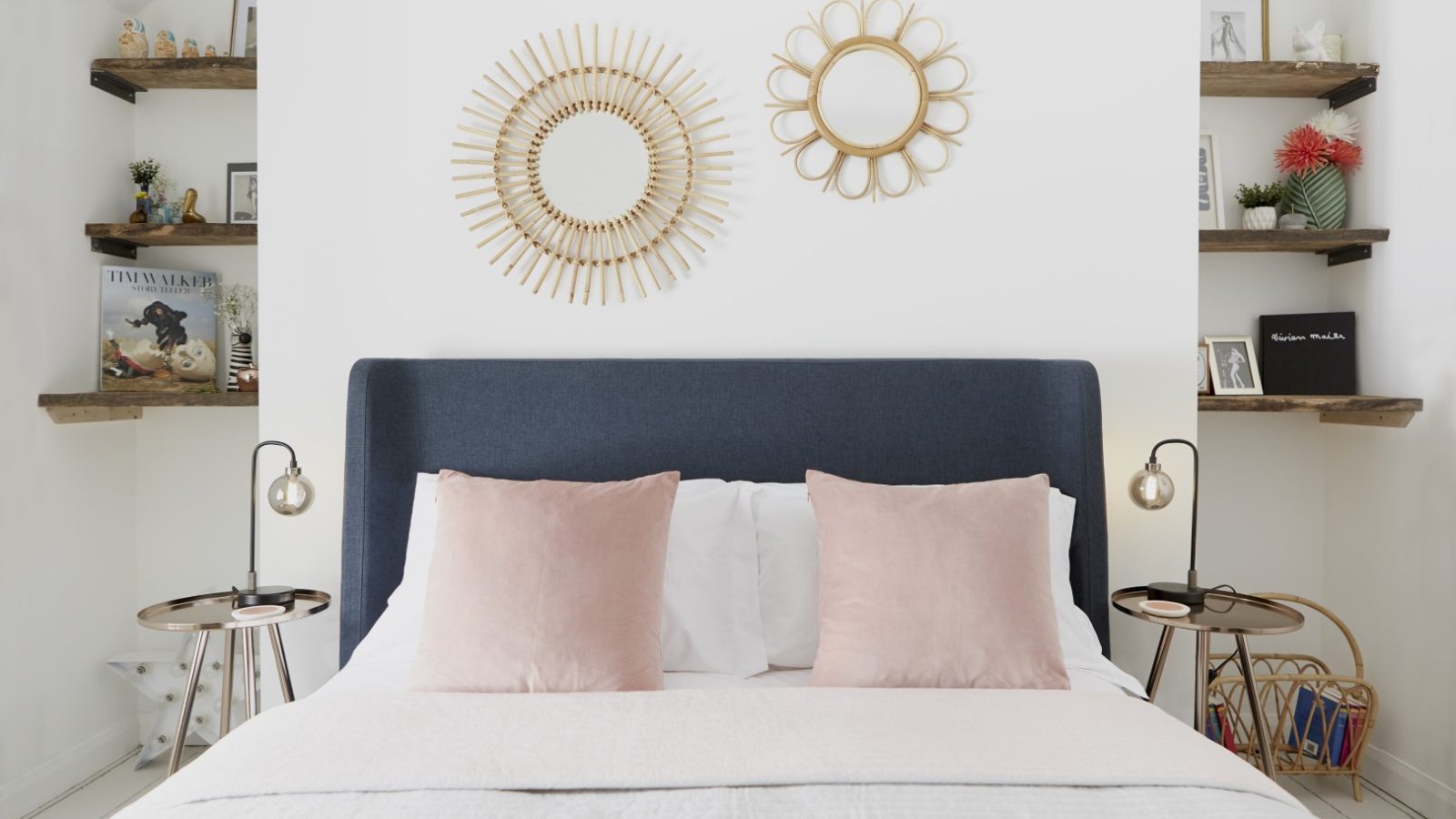 A neatly made bed with a navy-blue headboard and two pink cushions is centered against a white wall in Poachers Pocket Cottage. Above the bed hang two decorative round mirrors. Wooden shelves with books, photos, and decor items flank both sides of the room. Nightstands with lamps are on either side.