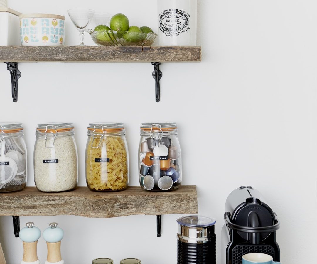 A kitchen setup at Poachers Pocket Cottage features wooden shelves stocked with jars containing various dry goods, spices, and cooking ingredients. Below the shelves, there's a counter with a coffee machine, a milk frother, a blue mug, and jars of spices neatly arranged.