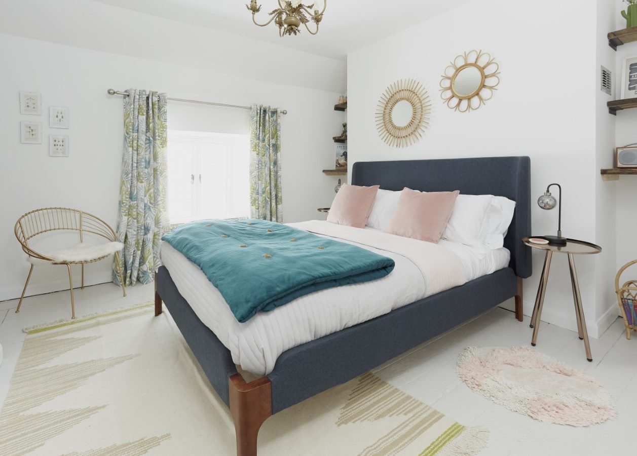 A modern bedroom with a large dark gray bed, white bedding, and a teal blanket. The room features white walls, two circular mirrors, tropical patterned curtains, a round side table, and a gold chandelier. A cozy chair is placed in the Poacher's Pocket corner near the window.