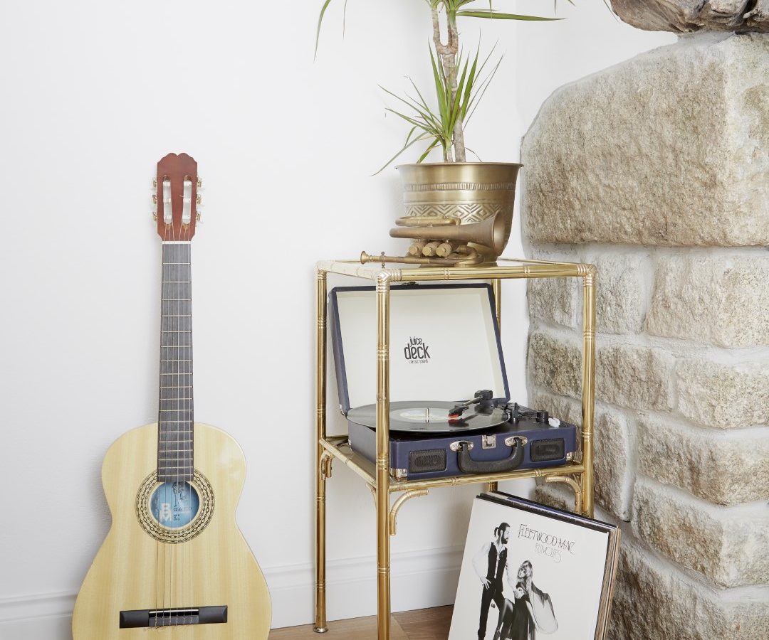 A cozy corner in Poachers Pocket Cottage features an acoustic guitar leaning against a white wall and a record player on a small gold and glass table. A potted plant sits on top of the table, with a vinyl record featuring Fleetwood Mac’s 