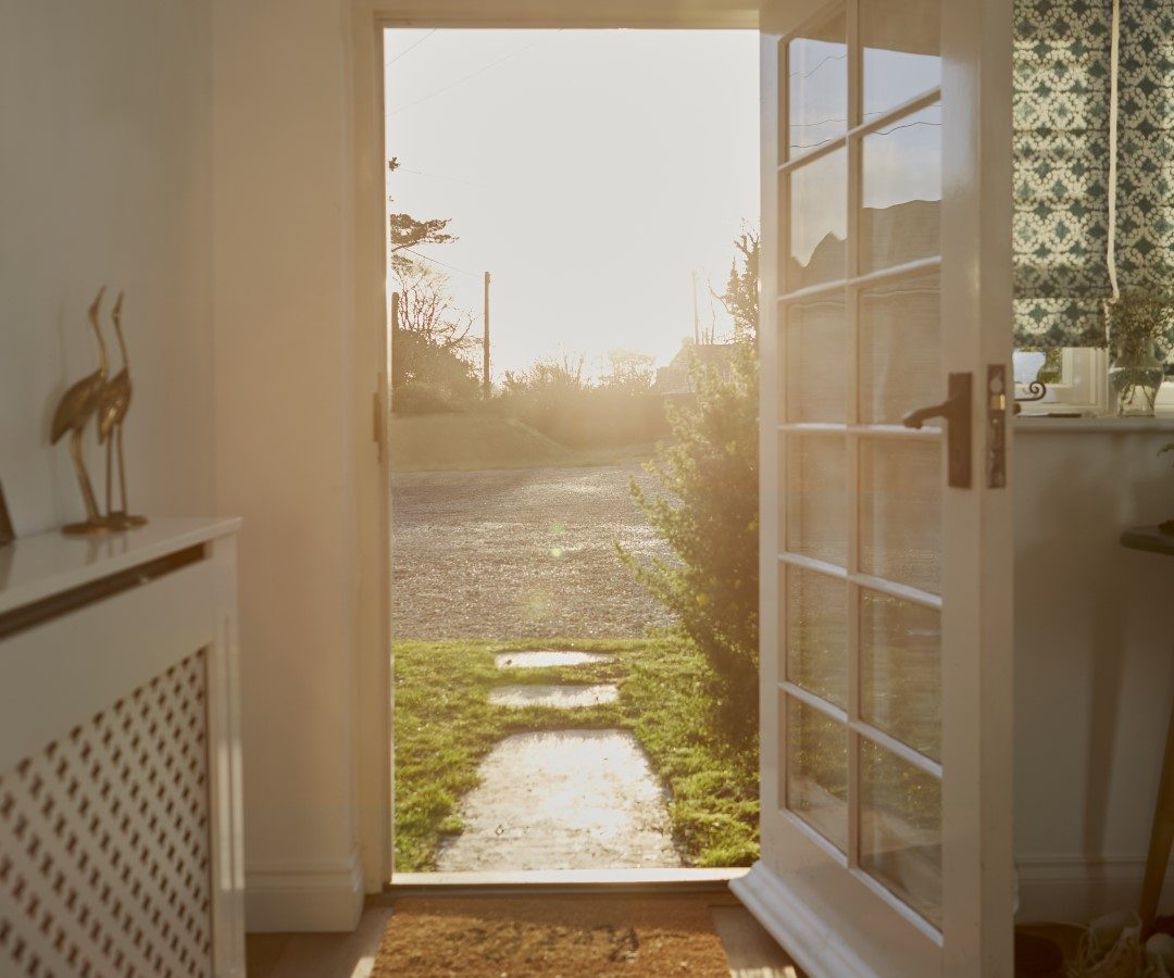 A door inside Poachers Pocket Cottage is open, revealing a sunlit pathway leading outside. The light from the sun illuminates the interior, showcasing a radiator cover with decorative bird statues on the left and a window with a patterned curtain on the right.