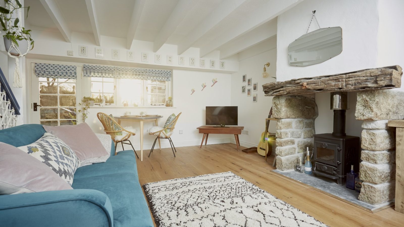 A cozy living room in the Poachers Pocket Cottage features a blue sofa, patterned cushions, and a white and black rug. A stone fireplace with a log burner warms the space, while a TV on a wooden stand provides entertainment. A small dining table sits by the window, white wooden beams accent the ceiling, and plants add greenery.