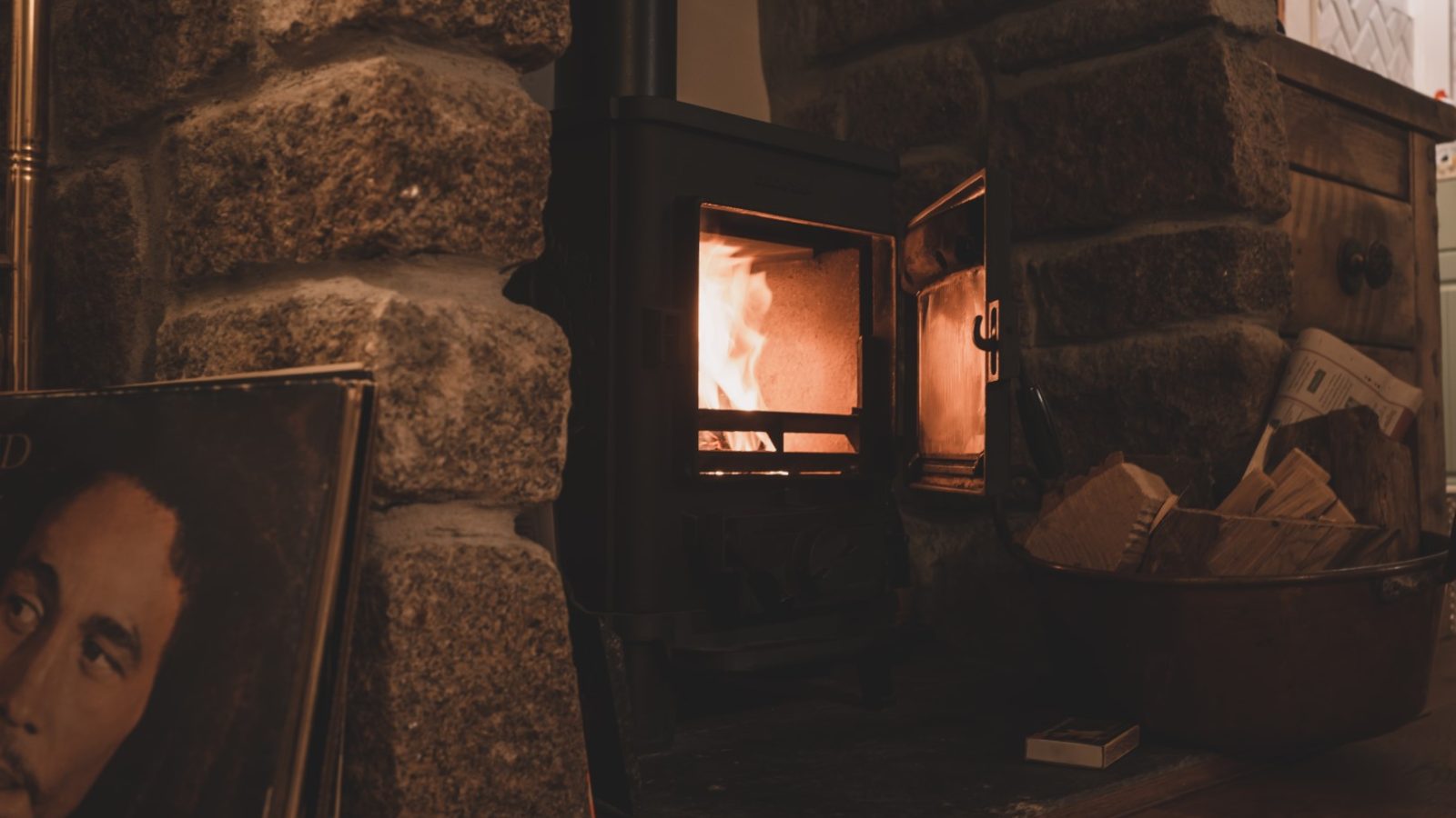 A cozy scene featuring a lit wood-burning stove set within a stone fireplace at the charming Poachers Pocket Cottage. The stove's door is slightly open, showing the glow of the fire inside. To the side, there is a vinyl record cover and a basket filled with firewood and newspapers.