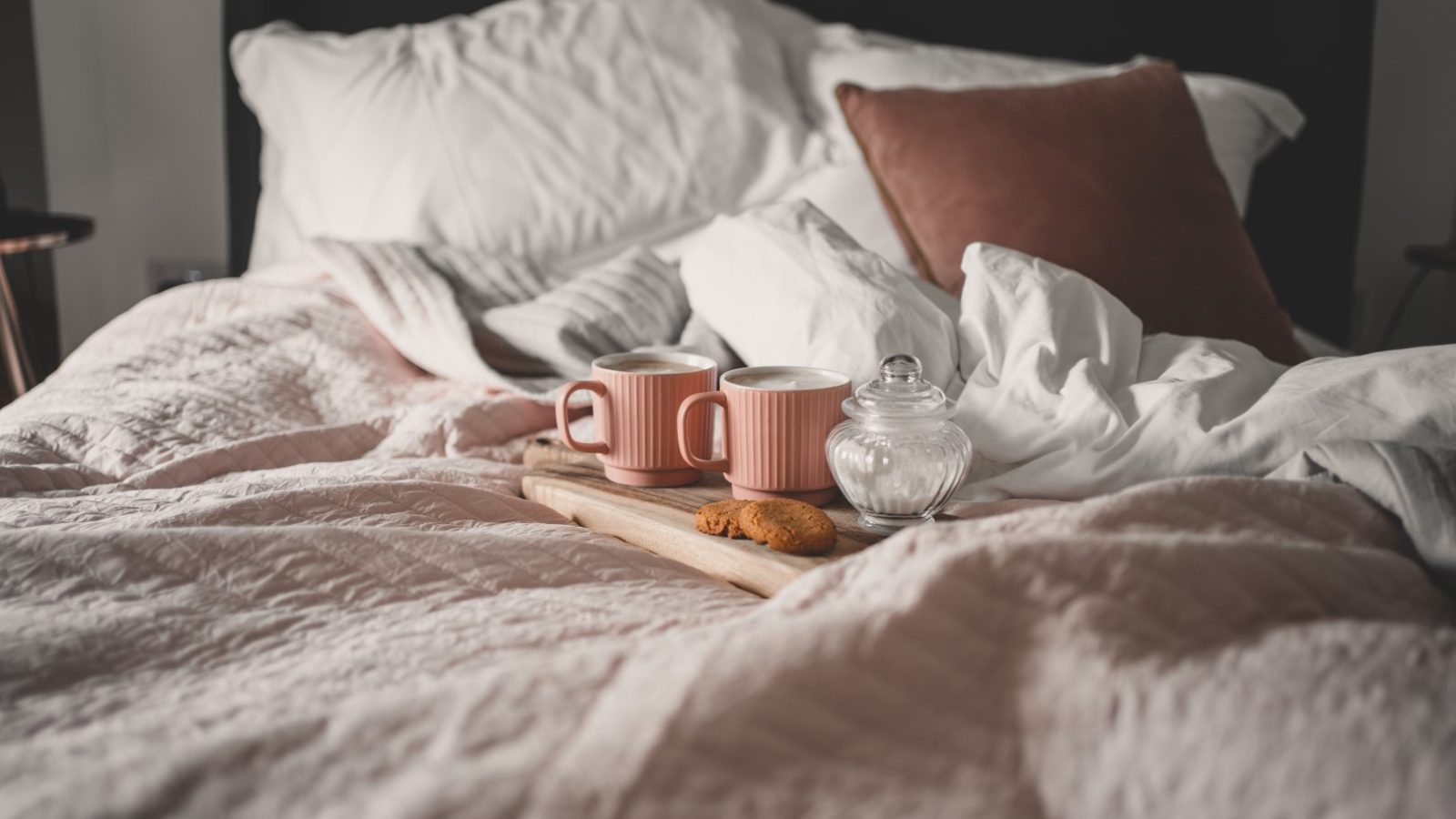 A cozy bed at Poachers Pocket Cottage with white and pink blankets and pillows, featuring a wooden tray holding two pink mugs, a glass jar, and a couple of biscuits. The scene exudes comfort and relaxation, with natural lighting enhancing the soft, inviting atmosphere.