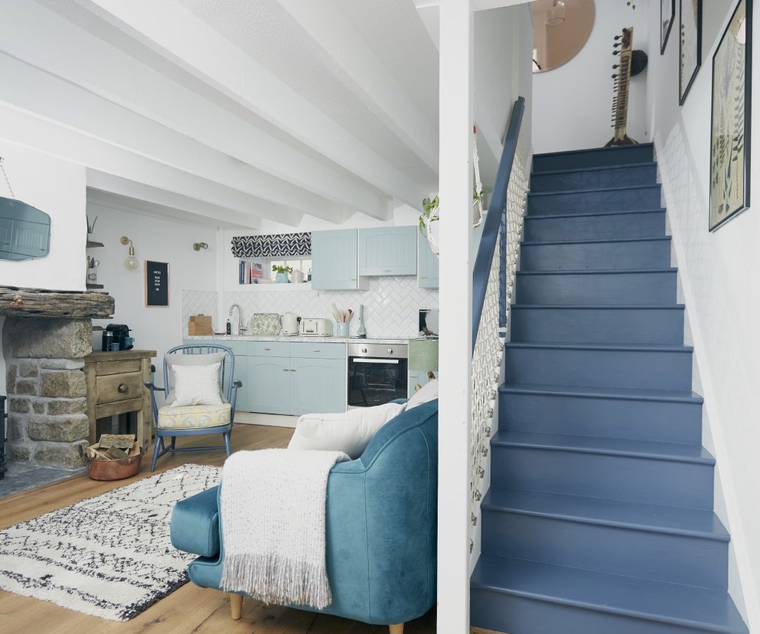 A cozy living space with a modern rustic theme at Poachers Pocket Cottage. Blue wooden staircase on the right, leading upstairs. On the left, a living area with a blue armchair, patterned rug, and stone fireplace. The kitchen with light blue cabinets is visible in the background.