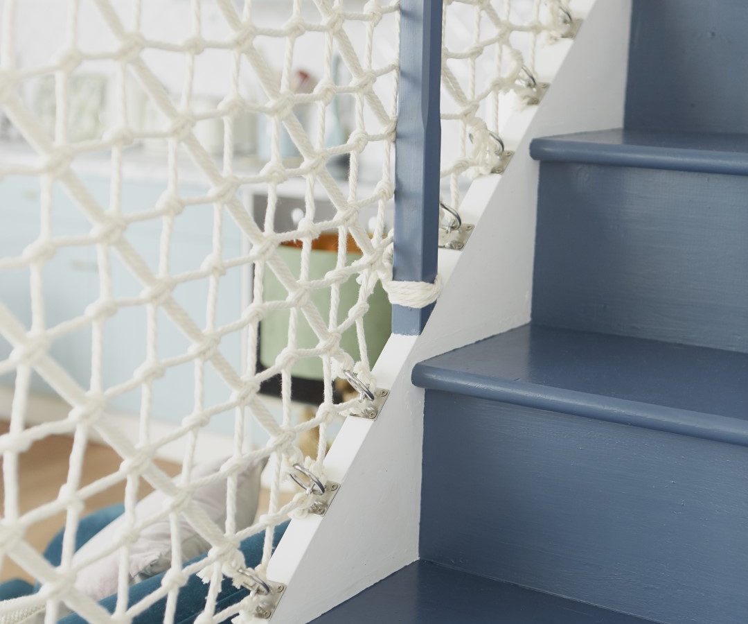 A cottage-style staircase with dark blue steps and white risers, featuring a railing made of white nautical netting. The background includes a light interior with pastel colors and soft lighting.