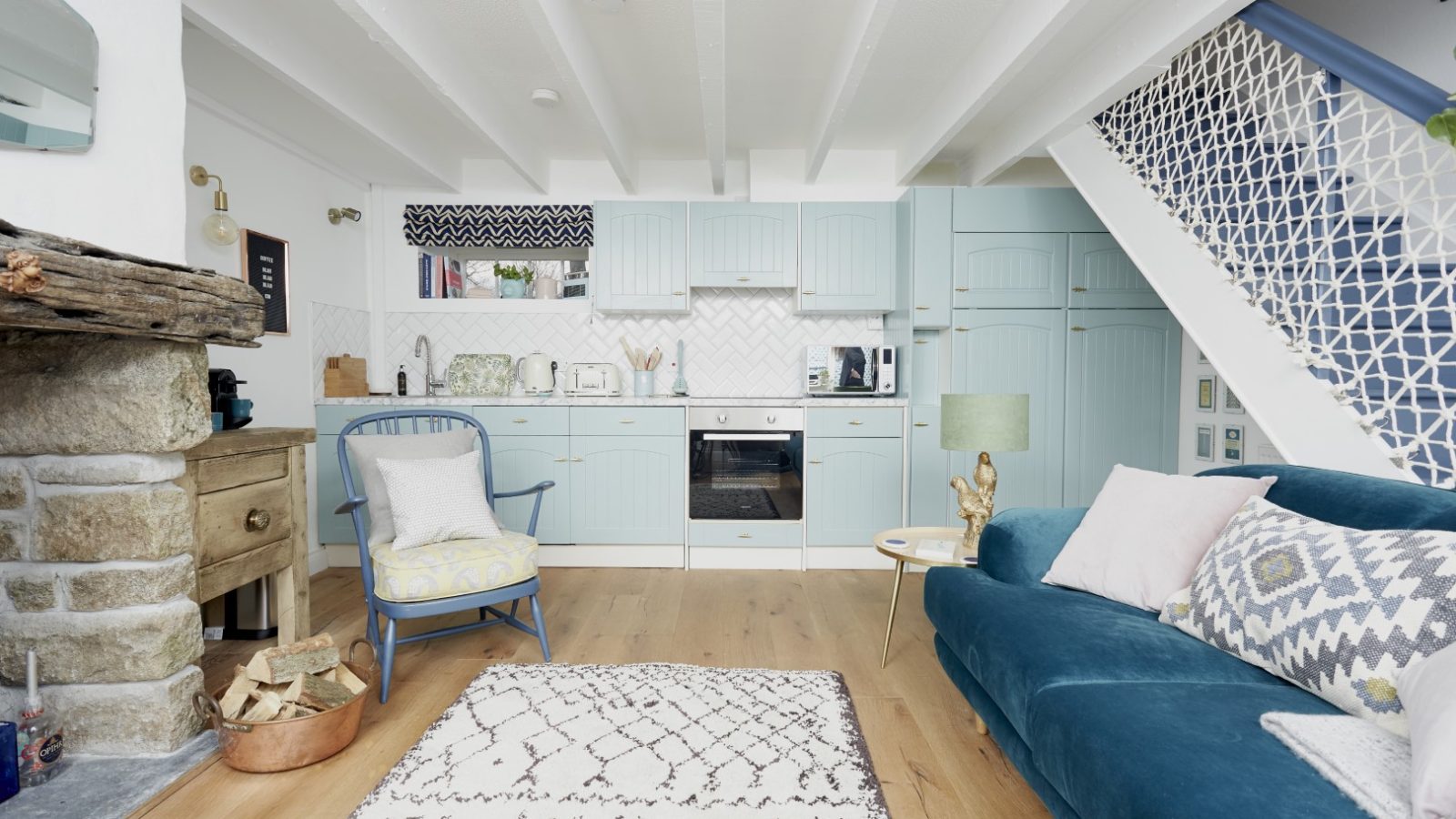 A cozy, rustic kitchen and living room at Poachers Pocket Cottage features light blue cabinets, wooden countertops, and a modern oven. A blue sofa with mixed cushions, a patterned rug, and a wooden chair with a cushion enhance the space. There's a decorative fireplace and a white staircase with lattice design.