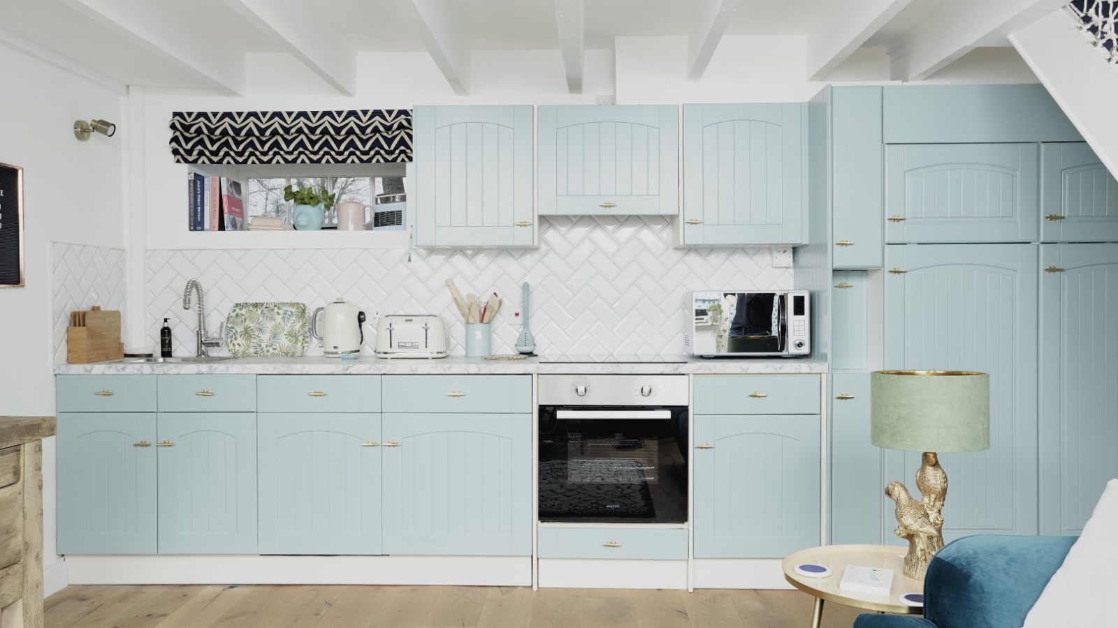 A modern kitchen with light blue cabinets, white countertops, and a herringbone tile backsplash evokes a cozy cottage charm. Appliances including a toaster, kettle, and microwave are on the counters. A green lamp sits on a side table near a dark blue chair in the foreground.