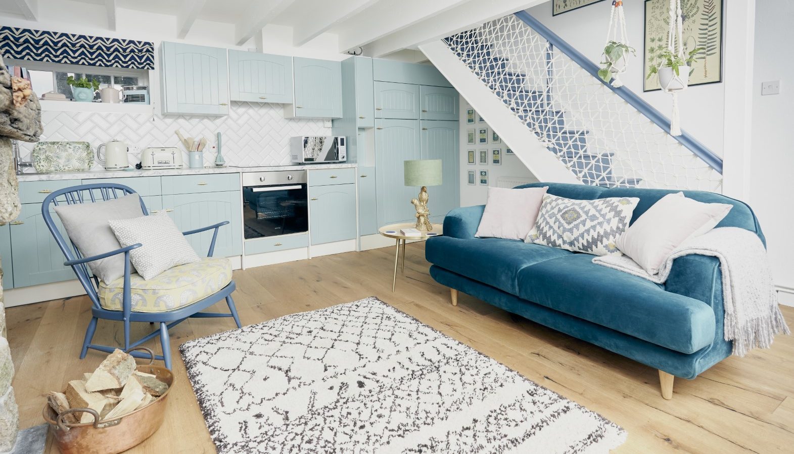 A cosy, modern living room and kitchen in Poachers Pocket Cottage features pastel blue cabinets, a teal sofa with cushions, and a white rug with black patterns. There's a rocking chair with cushions, plants hanging by the staircase, and wood flooring. The space has a bright, airy feel.