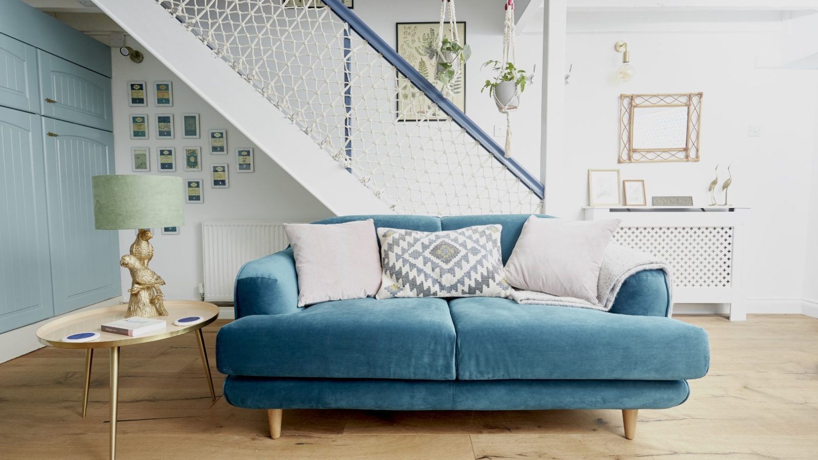 A modern living room at Poachers Pocket Cottage features a teal sofa adorned with assorted throw pillows. Behind the sofa is a wooden staircase with white netting. A small round table with a green lamp and figurine sits to the left. The space is decorated with framed photos and a console table.