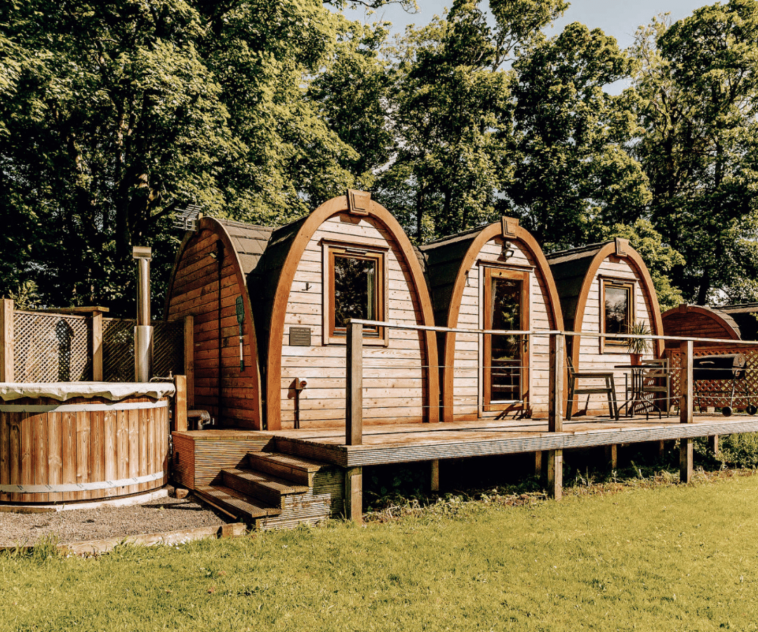 Wooden cabin pods with arched roofs are nestled among lush green trees. A wooden deck connects the pods, with a small outdoor seating area. To the left of the cabins is a wooden hot tub, where sunlight filters through blossoming leaves.