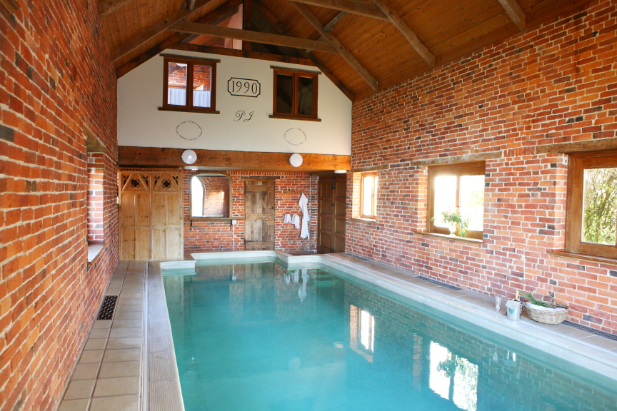 An indoor swimming pool with clear blue water is set inside a rustic room with exposed brick walls and wooden beams, reminiscent of Pennard Farm Cottages. Large windows on the right allow natural light to flood the space, and a sign with 