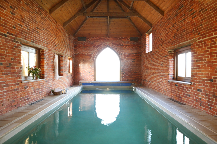 An indoor swimming pool with a rustic brick interior, wooden beam ceiling, and arched windows at Pennard Farm Cottages. The pool area features potted plants and ample natural light shining through the central arched window at the back.